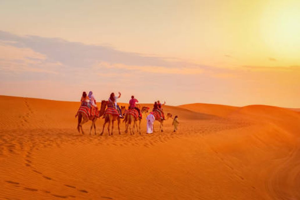 Caravan with group of tourists riding camels through Dubai desert during safari adventure, People exploring sand dunes at sunset in UAE