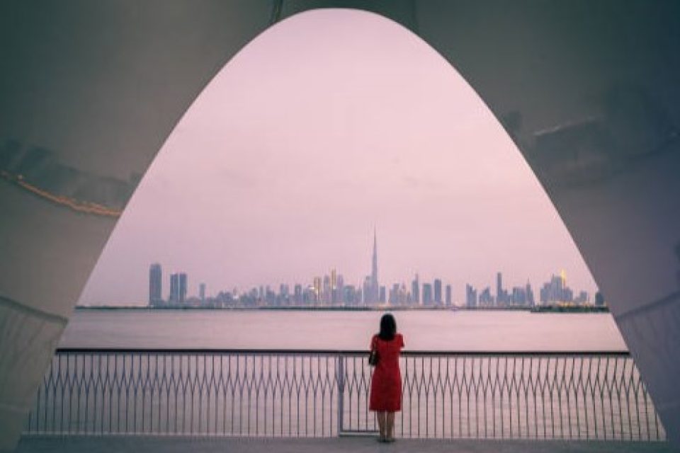 Woman standing enchanted, gazing at the breathtaking Dubai skyline from Creek Harbour. The city comes alive with twinkling lights against the dusky sky, creating a scene of tranquil beauty and urban allure